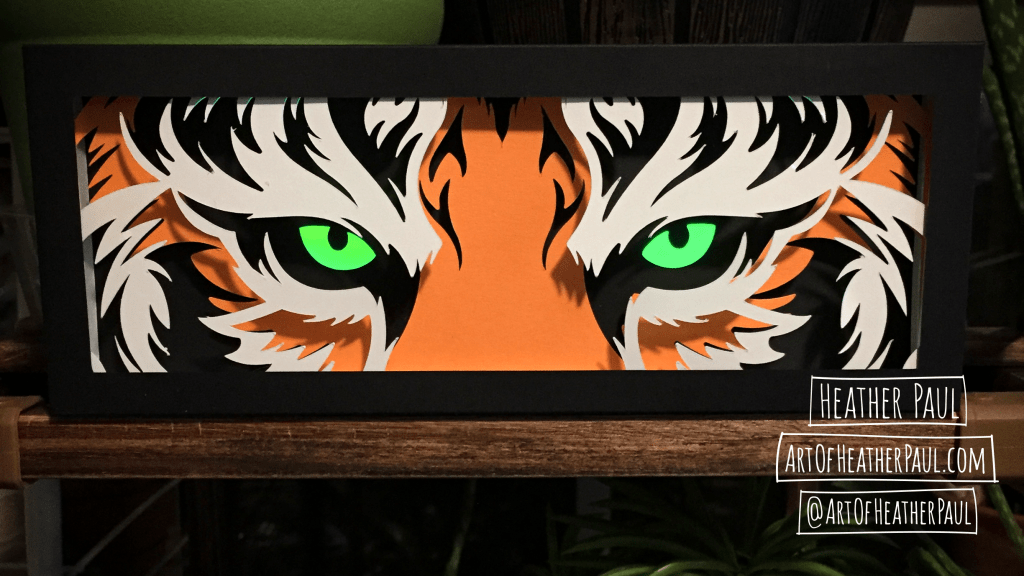 Photo of a back lit shadow box made of layers of cut paper making the image of tiger eyes (which are glowing) in full light