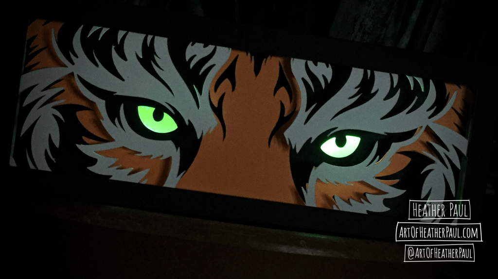 Photo of a back lit shadow box made of layers of cut paper making the image of tiger eyes (which are glowing) in dim light